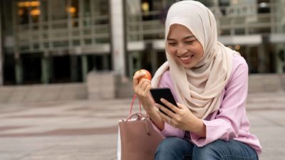 A young woman wearing an Islamic tradition headscarf and jeans sits in an urban environment with an apple in one hand and her phone in the other with a smile on her face.