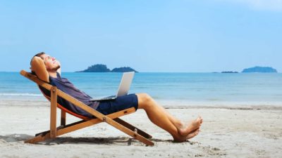 An ASX dividend investor lies back in a deck chair with his hands behind his head on a quiet and beautiful beach with blue sky and water in the background.