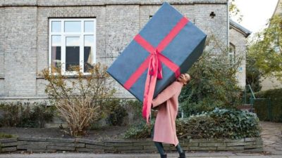A woman walks along the street holding an oversized box wrapped as a gift.
