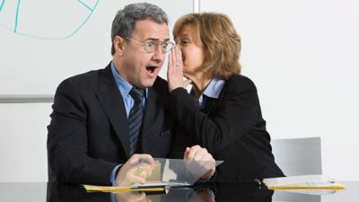 Businesswoman whispering in male colleague's ear as he looks surprised