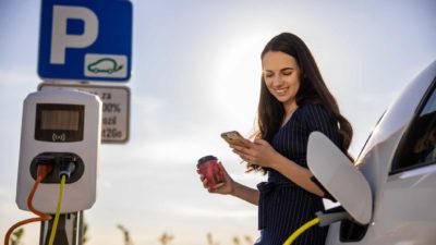a woman smiles as she checks her phone in one hand with a takeaway coffee in the other as she charges her electric vehicle at a charging station.