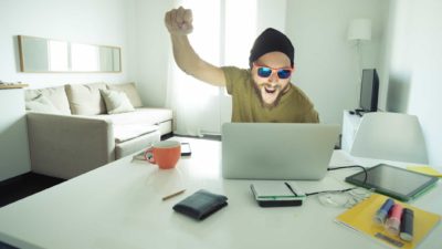 A man with a beard and wearing dark sunglasses and a beanie head covering raises a fist in happy celebration as he sits at is computer in a home environment.