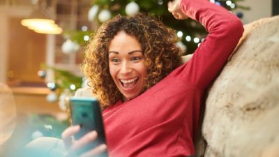 a woman raises her arm in celebration while looking at her mobile phone on her sofa at home feeling excited about the WiseTech share price rise