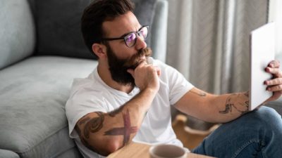 A hipster-looking man with bushy beard and multiple arm tattoos sits on the floor against a sofa reading a tablet with his hand on his chin as though he is deep in thought.