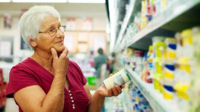 a woman ponders products on a supermarket shelf while holding a tin in one hand and holding her chin with the other.