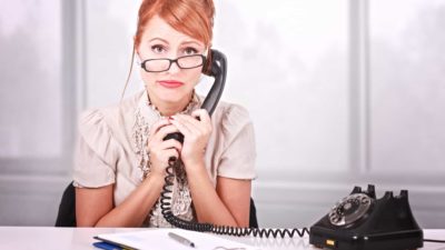 A woman holds an old fashioned telephone ear piece to her ear while looking unhappy sitting at a desk with her glasses crooked on her nose and a deflated expression on her face.