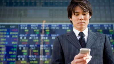 stock market news, person checks phone in front of electronic stock exchange boad