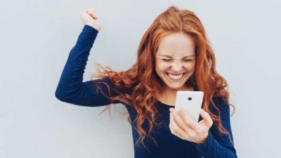 A woman with strawberry blonde hair has a huge smile on her face and fist pumps the air having seen good news on her phone.