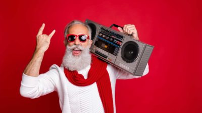 A cool older dude with a big white beard and wearing a red scarf holds a boombox stereo on his shoulder and makes rock'n'roll devil fingers with his other hand.