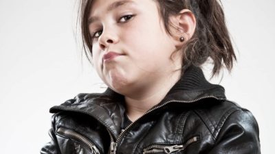 A young girl wearing a leather jacket with zips looks up slyly with one eyebrow raised.