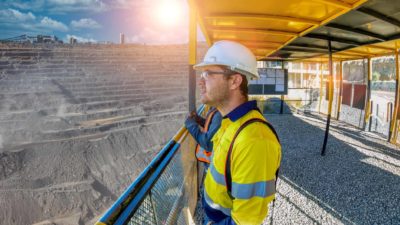 A mining worker wearing a white hardhat and a high vis vest stands on a platform overlooking a huge mine, thinking about what comes next.