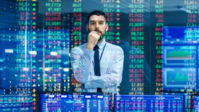 A male ASX 200 broker wearing a blue shirt and black tie holds one hand to his chin with the other arm crossed across his body as he watches stock prices on a digital screen while deep in thought