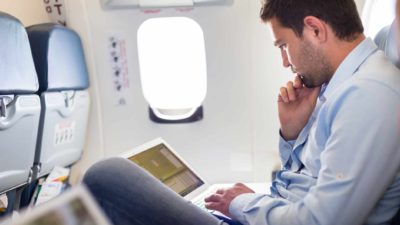 Man sitting in a plane seat works on his laptop.