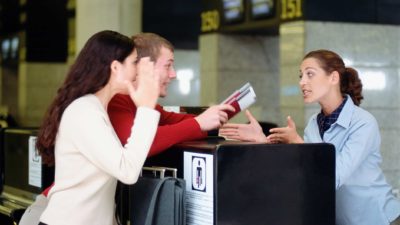 a couple at an airline ticket counter have an angry exchange with the employee behind the counter. She is leaning forward in an aggressive manner as they hold a paper ticket in their hands.