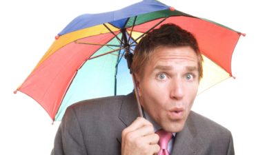 a man wearing a suit and holding a colourful umbrella over his head purses his lips as though he has just found out some interesting news.