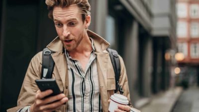 A cool young man walking in a laneway holding a takeaway coffee in one hand and his phone in the other reacts with surprise as he reads the latest news on his mobile phone