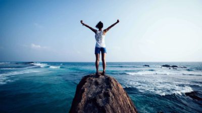 A beautiful ocean vista is shown with a woman whose back is to the camera holding her arms up in triumph as she stands at the top of a rock feeling thrilled that ASX 200 shares are reaching multi-year high prices today